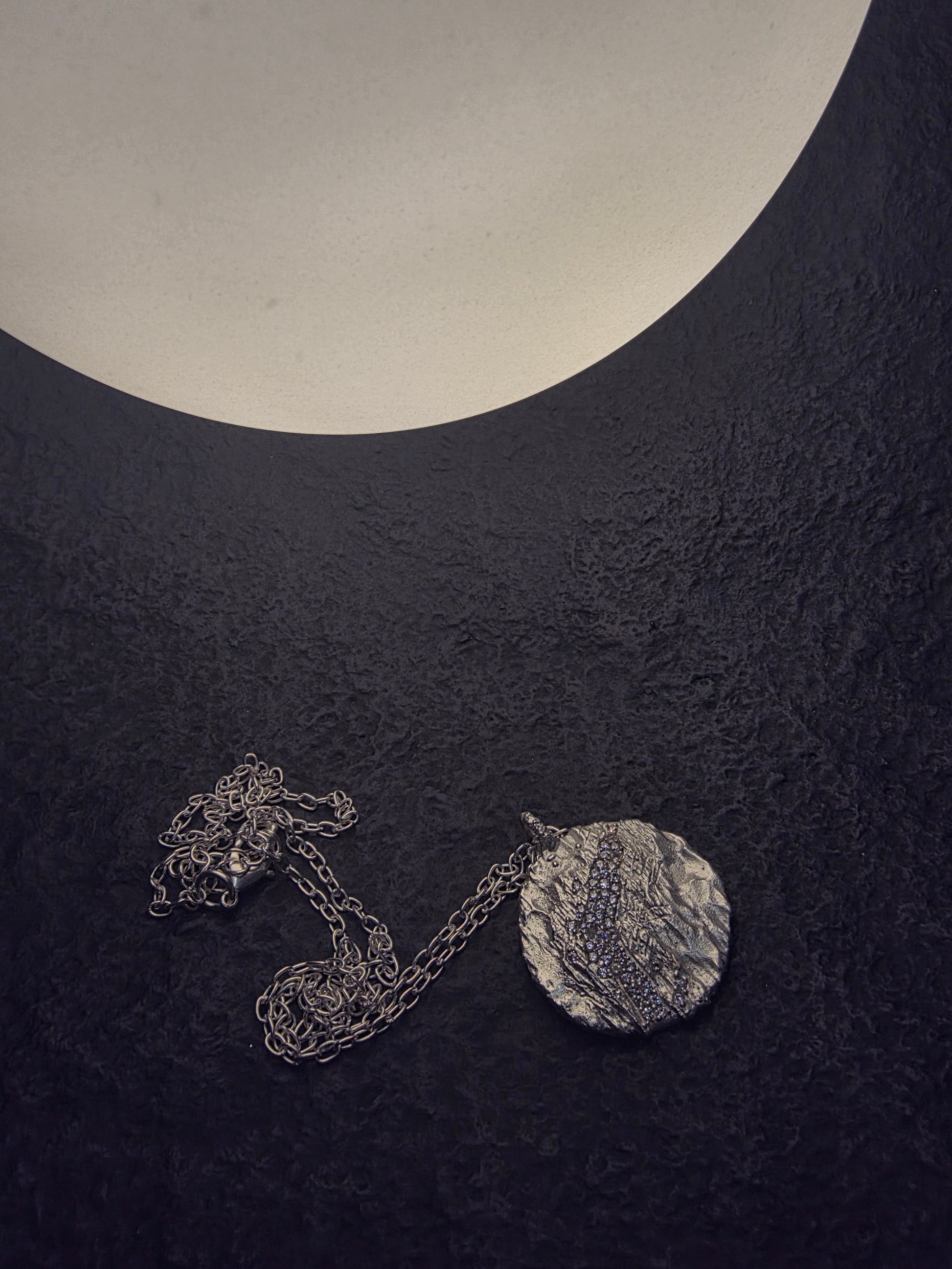Near Earth Object | Silver necklace with a round pendant on a dark textured surface