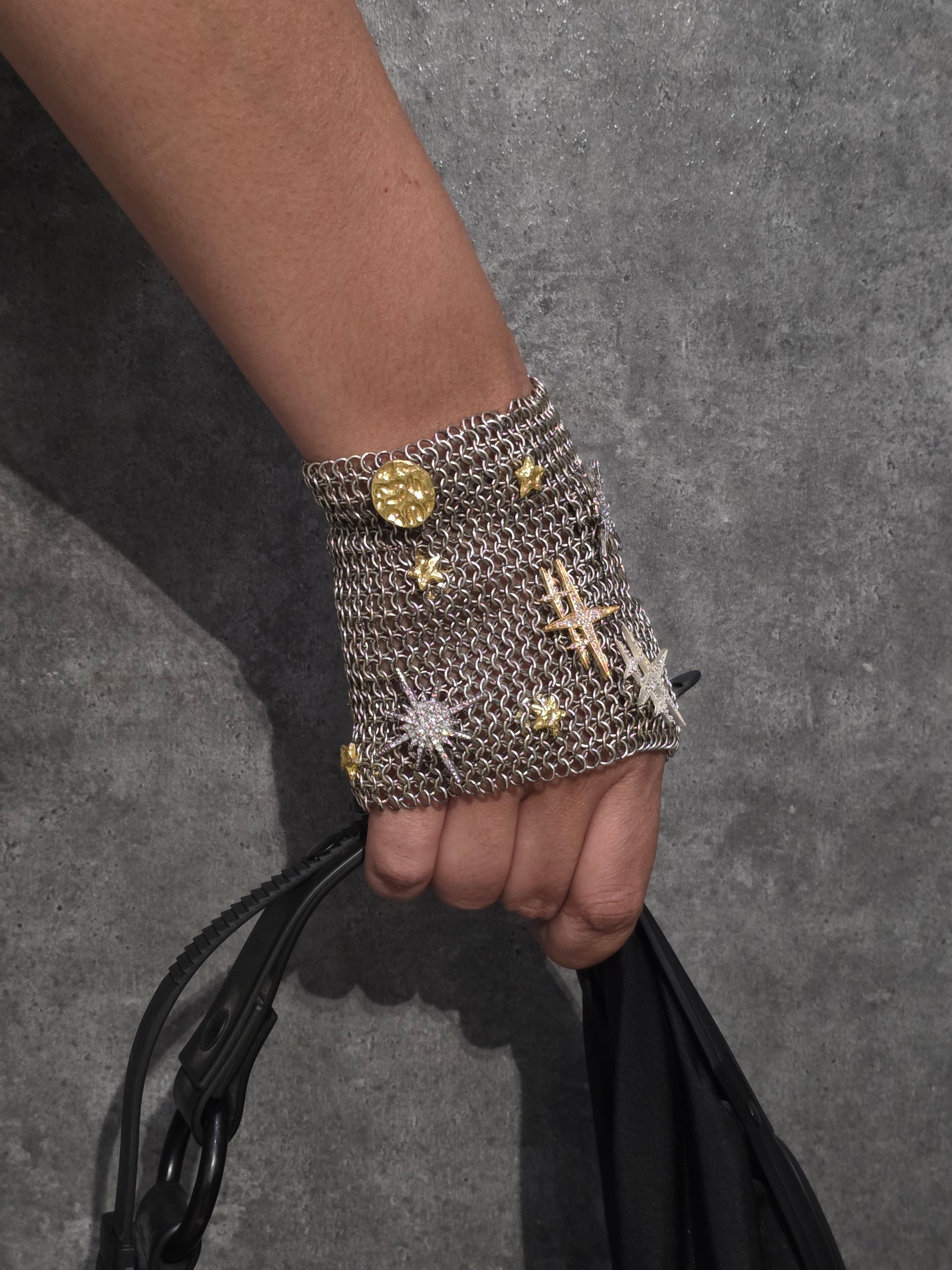 Near Earth Object | A person's hand wearing a silver chain bracelet with gold accents, holding a black bag.