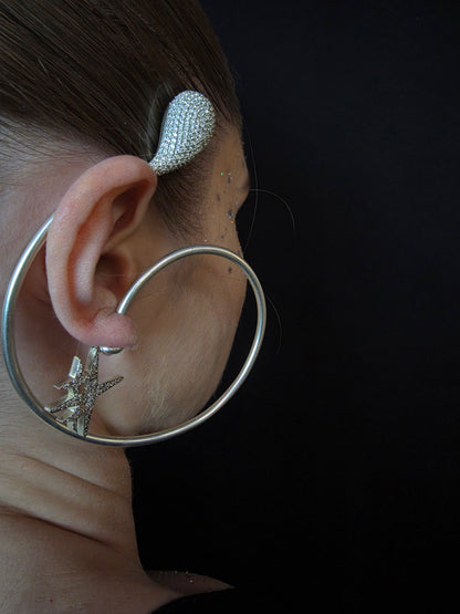 Near Earth Object | Close-up of an ear with silver hoop earrings and a decorative earring against a dark background