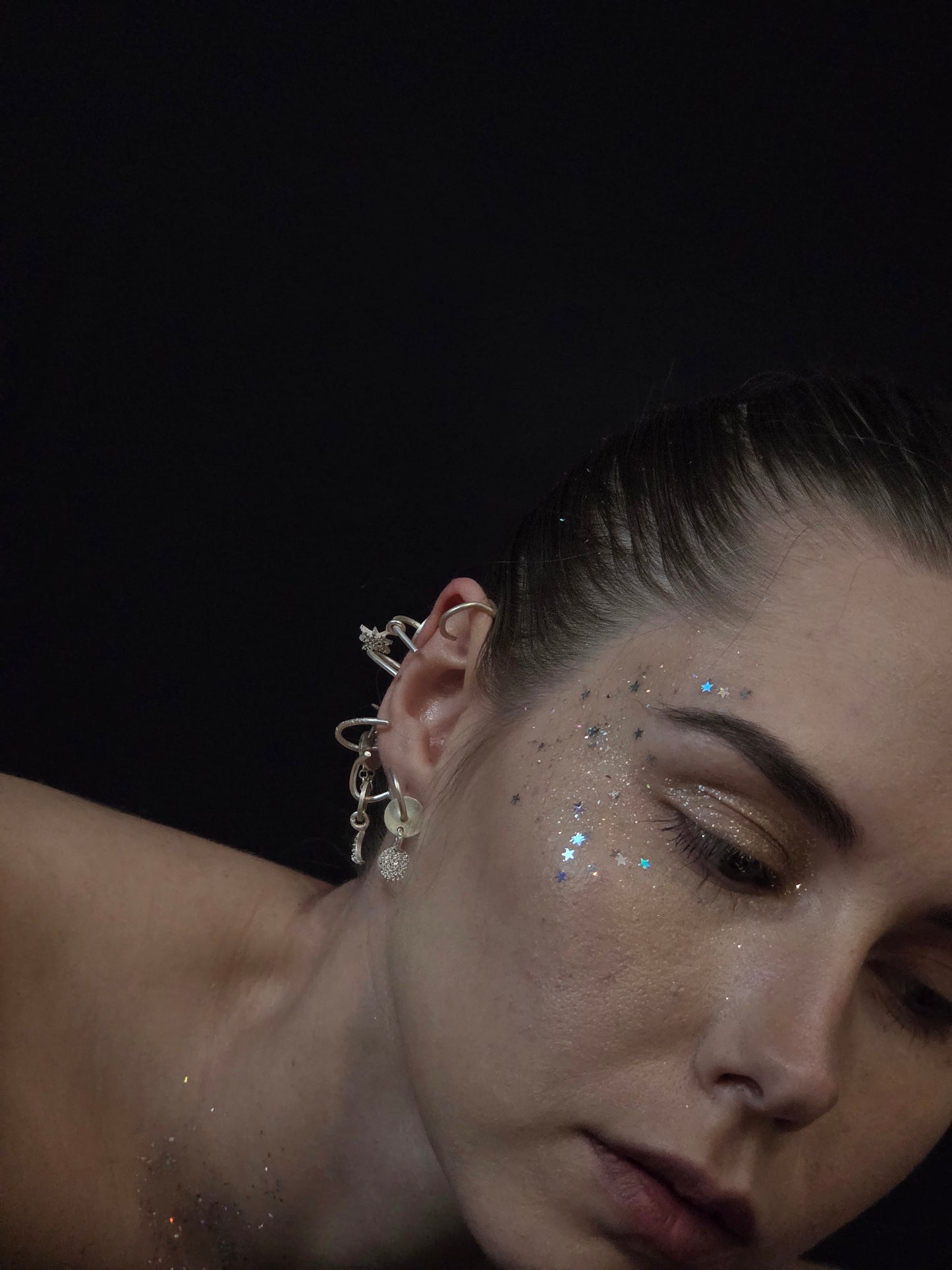 Near Earth Object | Close-up of a person with decorative earrings and face makeup against a dark background