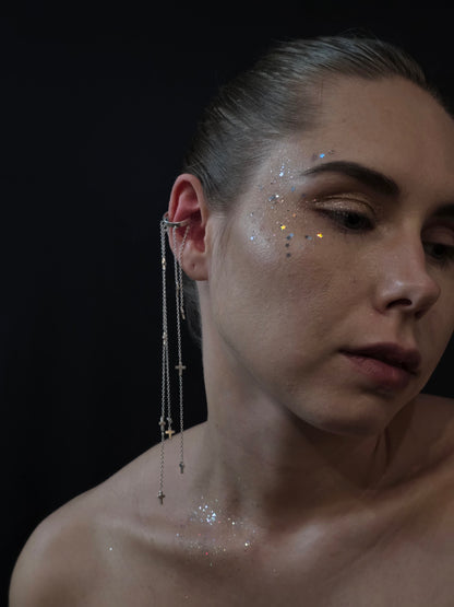 Near Earth Object Cross Cascade Cuff worn on model – dramatic silver multi-chain ear cuff with cross charms falling from ear to shoulder, paired with iridescent star glitter on cheek and décolletage