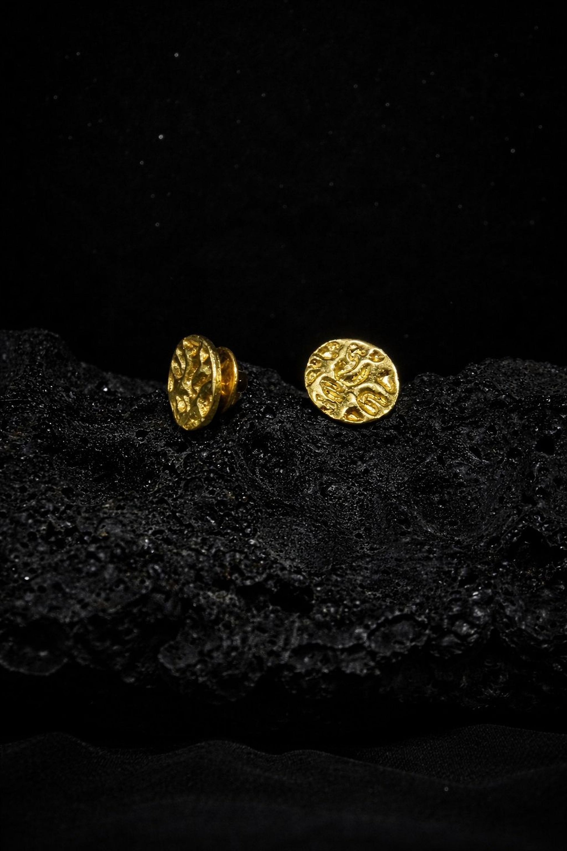 Two gold earrings with intricate designs are resting on a black surface, creating a striking contrast.
