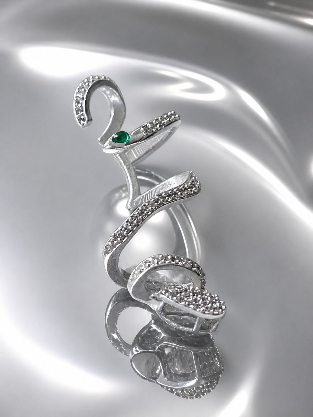 Near Earth Object | Silver snake-shaped brooch with a green gemstone on a reflective silver background