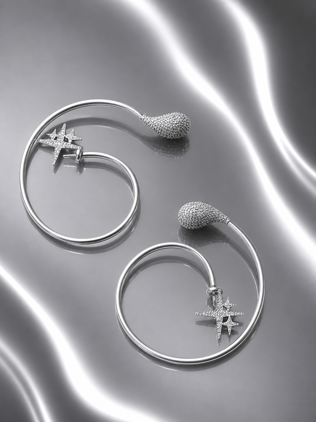 Near Earth Object | Silver hoop earrings with star and sphere designs on a reflective surface