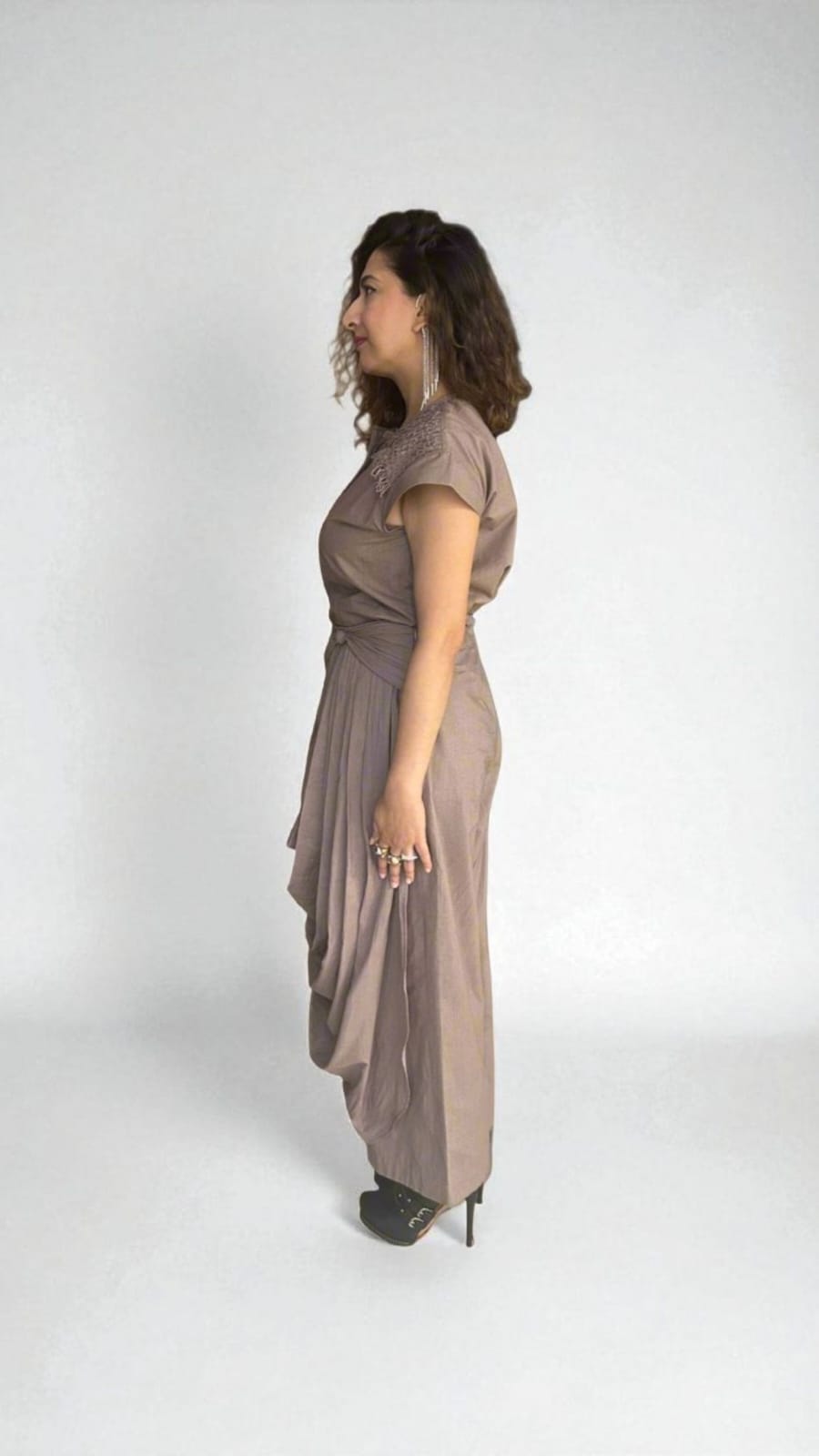 Crater Matter Set : Side profile of a woman wearing a taupe draped  Dress with pleated detailing at the waist and cascading fabric on the side, paired with black stiletto heels and statement rings, modern ethnic fusion  Dress with gathered pleats. | Women's  Dress | Pret Clothes | Asymmetrical dress | nEo Clothes.