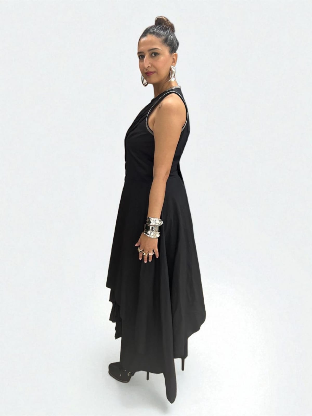 In side view Women wearing a modern black asymmetrical Jumpsuit with pleated detailing, The Jumpsuit has a sleeveless design with metallic edging at the neckline. Accessorizes with bold silver jewelry including hand cuff, stacked rings, and hoop earrings, The overall style is a fusion of contemporary and ethnic fashion, giving off a confident, Trendy sleeveless Jumpsuit outfit for women with ethnic silver jewelry. | Women's Jumpsuit | Pret Clothes | Asymmetrical Jumpsuit | nEo clothes