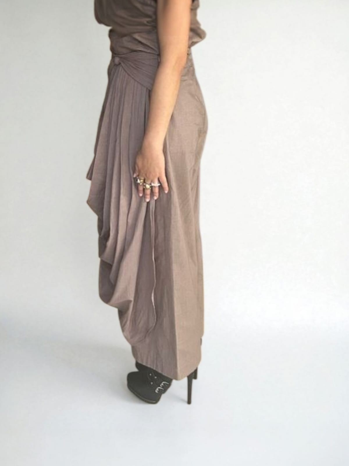 Side profile of a woman wearing a taupe draped  Pant with pleated detailing at the waist and cascading fabric on the side, paired with black stiletto heels and statement rings, modern ethnic fusion  Pant with gathered pleats. | Women's  Pant| Pret Clothes | Asymmetrical Pant | nEo Clothes.